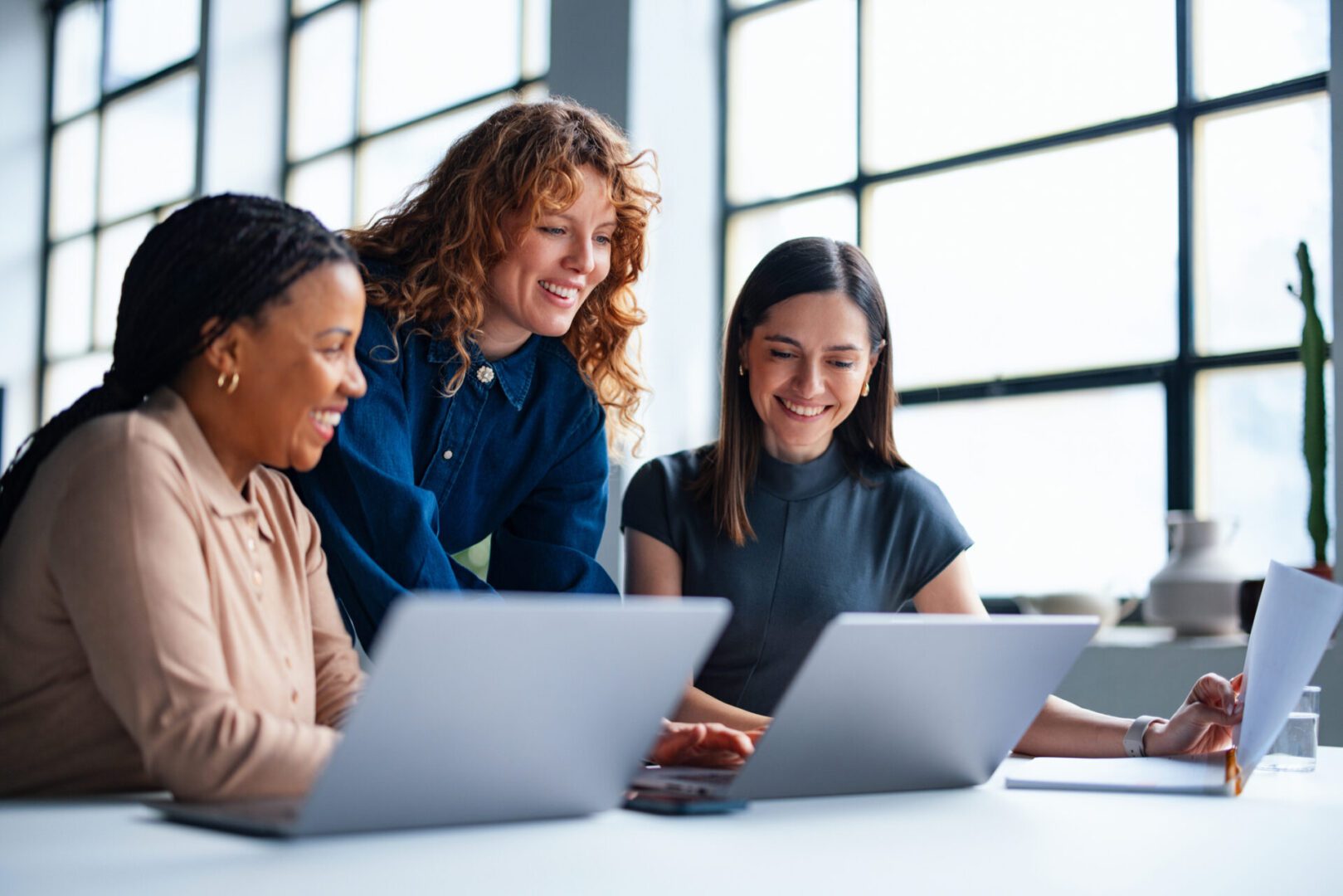 Three women collaborating on laptops in office.