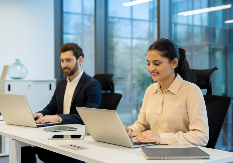 Two colleagues working on laptops in a modern office setting, teamwork concept. The scene shows collaboration and productivity.