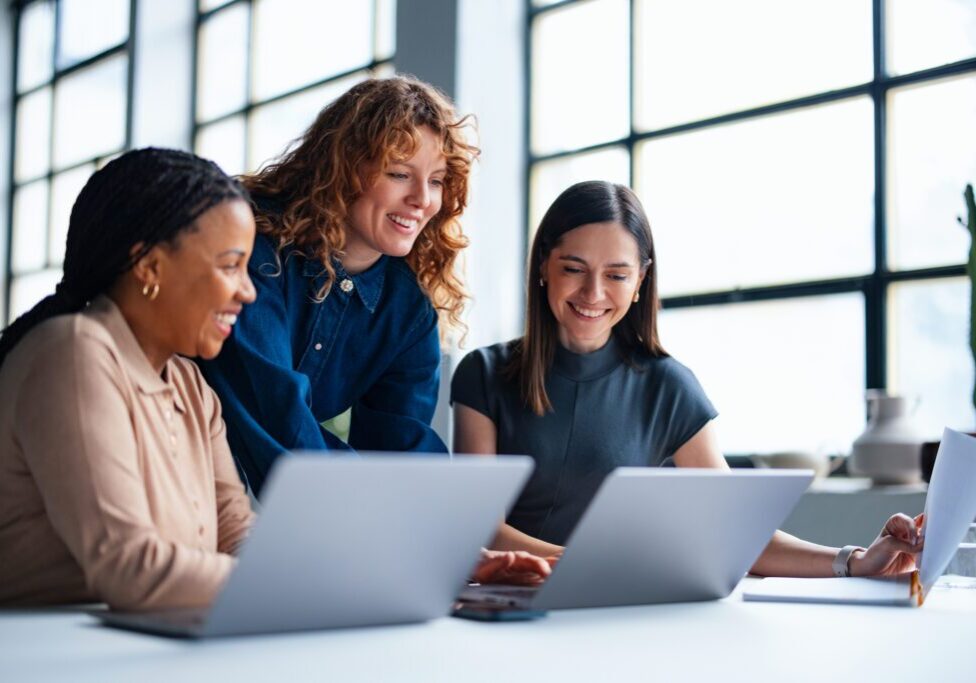 Three women collaborating on laptops in office.